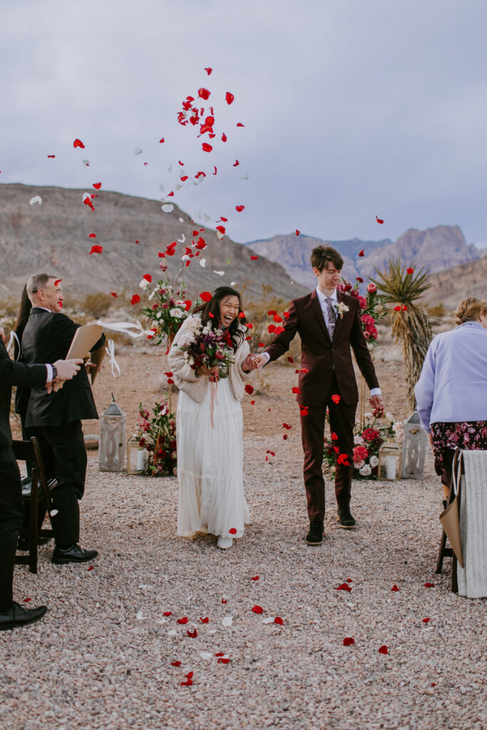 Flower petals are thrown above the bride and groom as they walk hand in hand as husband and wife.  The bride wears a V-neck silk top with a long shoulder cape and tulle flowy skirt. The groom wears a deep maroon suit with purple paisley tie. 
