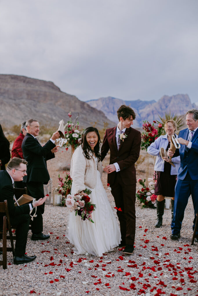 Love Unleashed: Colorful Las Vegas Elopement. Ceremony guests cheer as the bride and groom walk down the isle over red and pink flower petals.  The The bride wears a V-neck silk top with a long shoulder cape and tulle flowy skirt. The groom wears a deep maroon suit with purple paisley tie. 