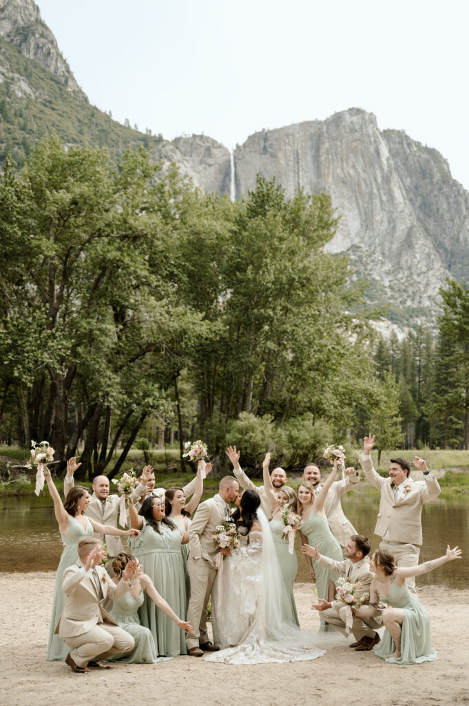 Bridesmaids and groomsmen in mint green and light tan suits and dresses, cheer excitedly as the bride and groom share a kiss. The dramatic wide angle photo shows Yosemite national park behind them. 