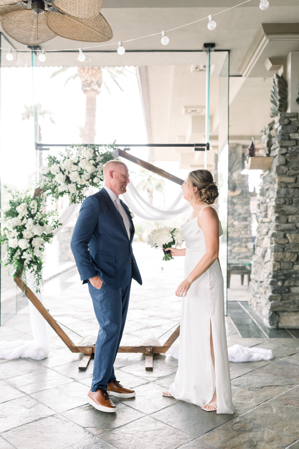 Groom in a navy blue suit standing in front of a octagon wooden back arch, looks at his bride for the first time. 