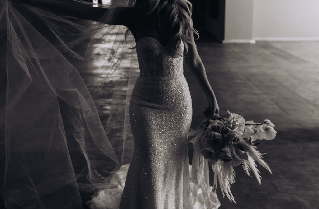 The Ultimate Guide to Different Types of Wedding Dresses: From Classic to Contemporary. Black and white photo of a fitted sleeveless sparkly  wedding dress. The bride holds her boho styled bouquet down at her side. 