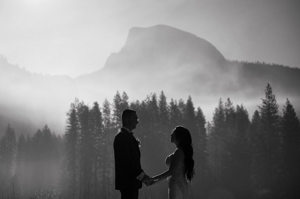 Unveiling the Magic: The Art of Styling a Themed Wedding black and white photo of the foggy mountains and pine trees. A bride and groom hold hands silhouetted against the trees. 