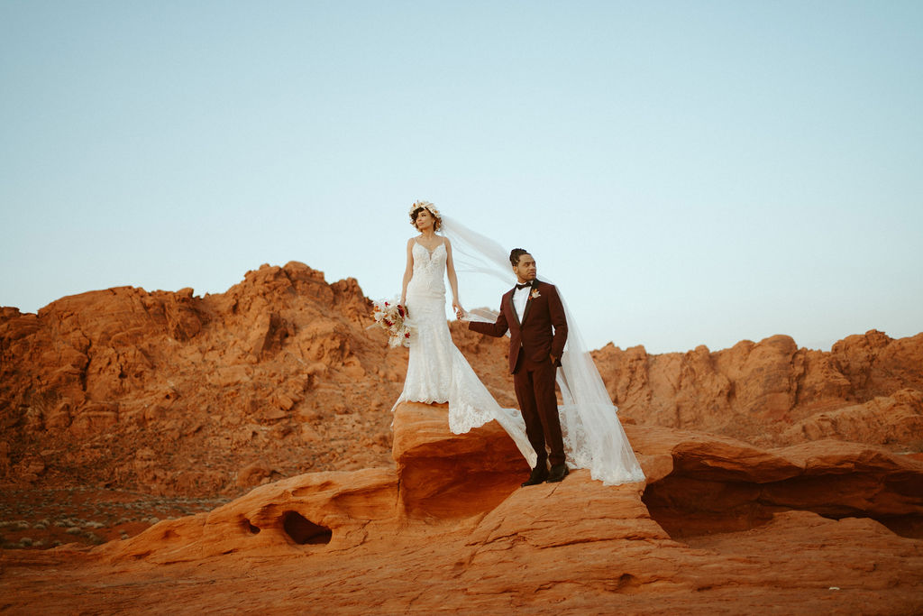 Fun & Creative Wedding Day Photo Poses and Ideas Bride and groom standing on top of a red rock. The bride weares a spaghetti strap lace fitted wedding gown. With a floral head piece and long veil flowing in the wind. The groom is wearing a deep rich maroon suit with black shoes and bowtie. 