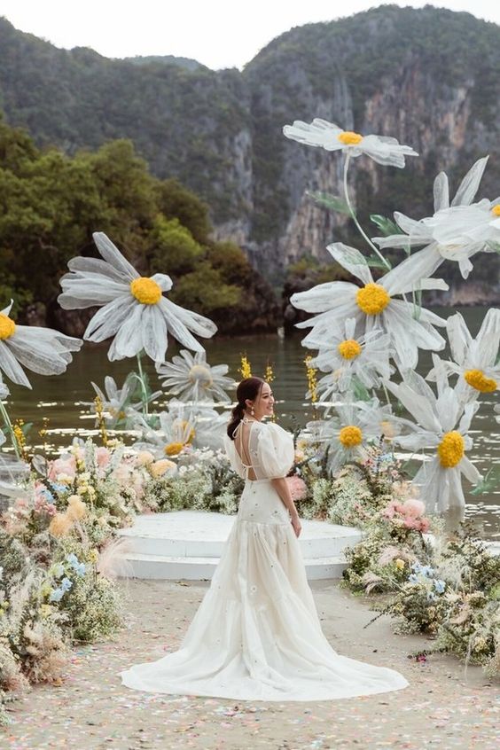 Unveiling the Magic: The Art of Styling a Themed Wedding. Bride with puffy white sleeves and open back stands in from of her ceremony backdrop with massive wire and tule white and yellow daisies. 