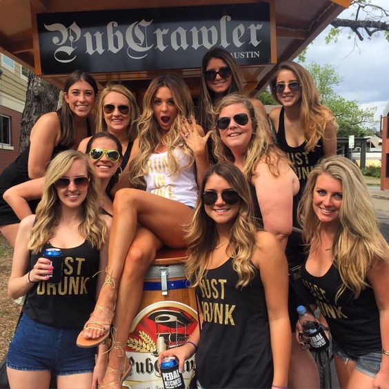 Bride sitting on a whisky barrel in a white tank top. Her bridesmaids are wearing black tank tops that say just drunk. A pub crawl sign is above them. 