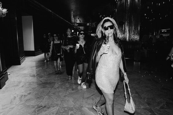 Black and white photo of bride leading her group of friends in Vegas. She wears sunglasses, viel, and white sparkle dress. Sipping on a drink as the bridal party is in all black behind her. 