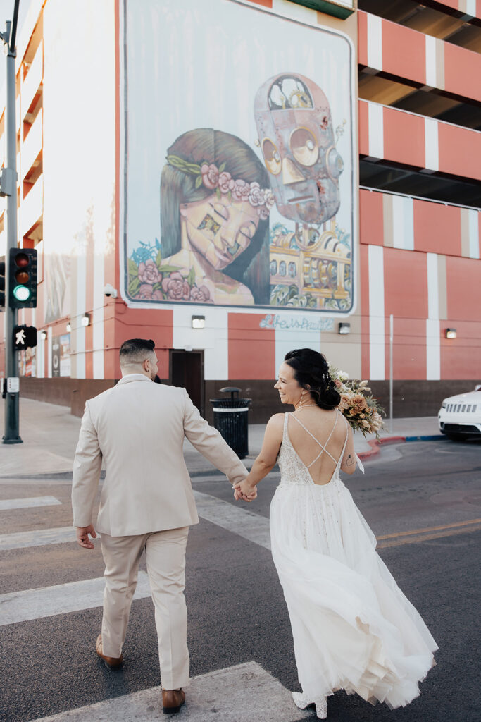 Bride and groom walk hand across a street walkway. A beautiful building mural is in the background. The groom wears a light tan suit. The brides wedding dress flows with an open back and sparkly boots underneath. 