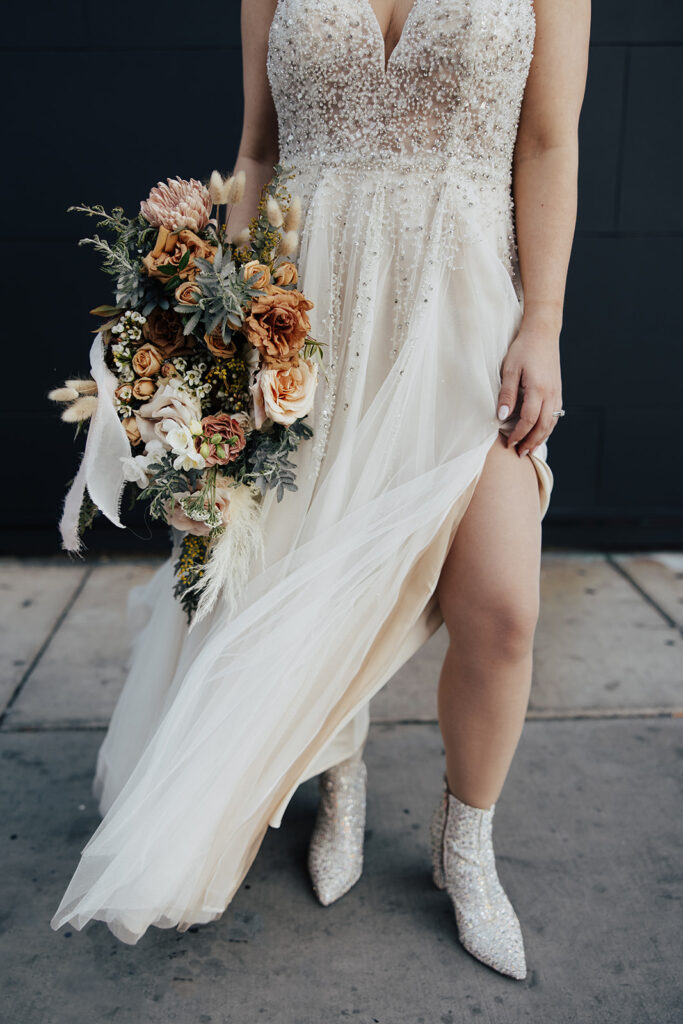 Bride wearing a vintages tea-length dress with glittering boots. Her sparkling tea-length wedding dress has a deep slit and v-neck neckline. Close up of the dress shows the intricate glittering details. She holds a sage green, soft orange, cream, and soft pink bridal bouquet. 