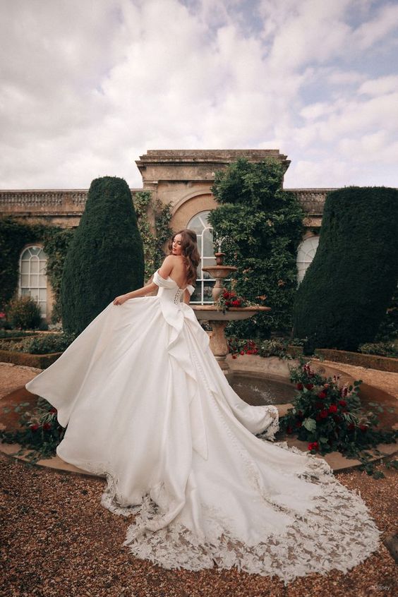 Couture off the shoulder sleeves bridal gown . Dramatic long train with lace. The bride stands in front of a historical building. 
