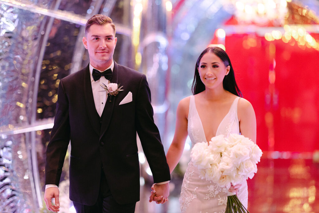 Couple walks in through a glass tunnel with hanging chandeliers and lights that look like water. The groom wears an fitted black tux. The bride is wearing a fitted deep V-neck mermaid bridal gown. She holds an all white rose bridal bouquet