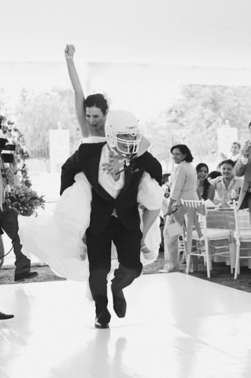 A black and white photo of a bride getting a piggy back ride from her groom. The groom wears a white football helmet as they run across their dance floor for their grand entrance. 