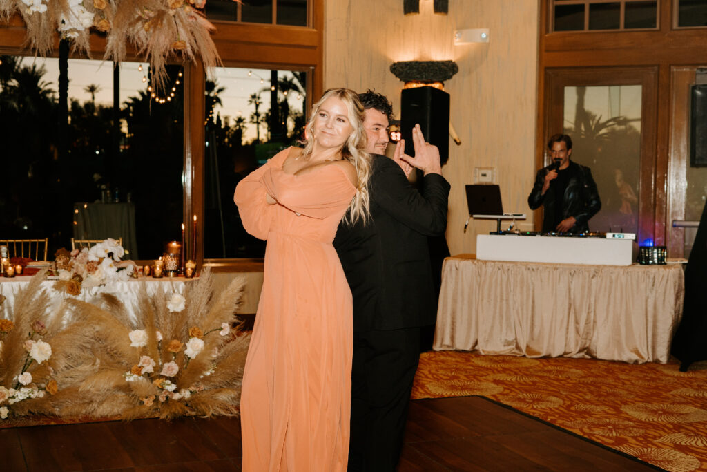 Bridesmaid and groomsman strike a pose for their part in the grand entrance of the reception. Teh bridesmaid is in a peach orange long sleeve dress. The groomsman is in a black suit. 
