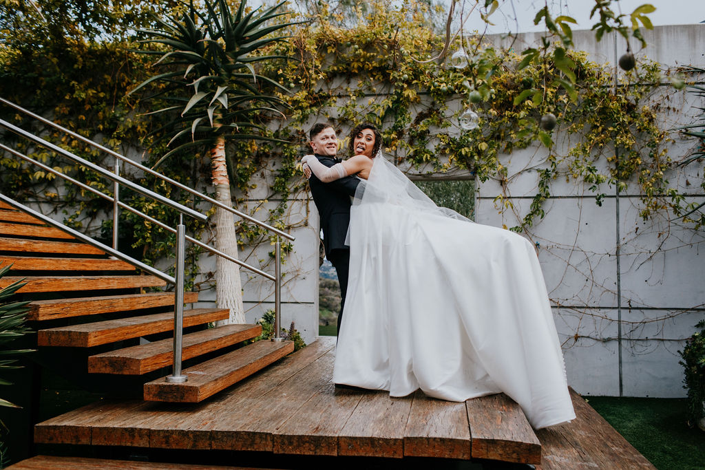Groom holding up bride outside modern venue with stairs and greenery 