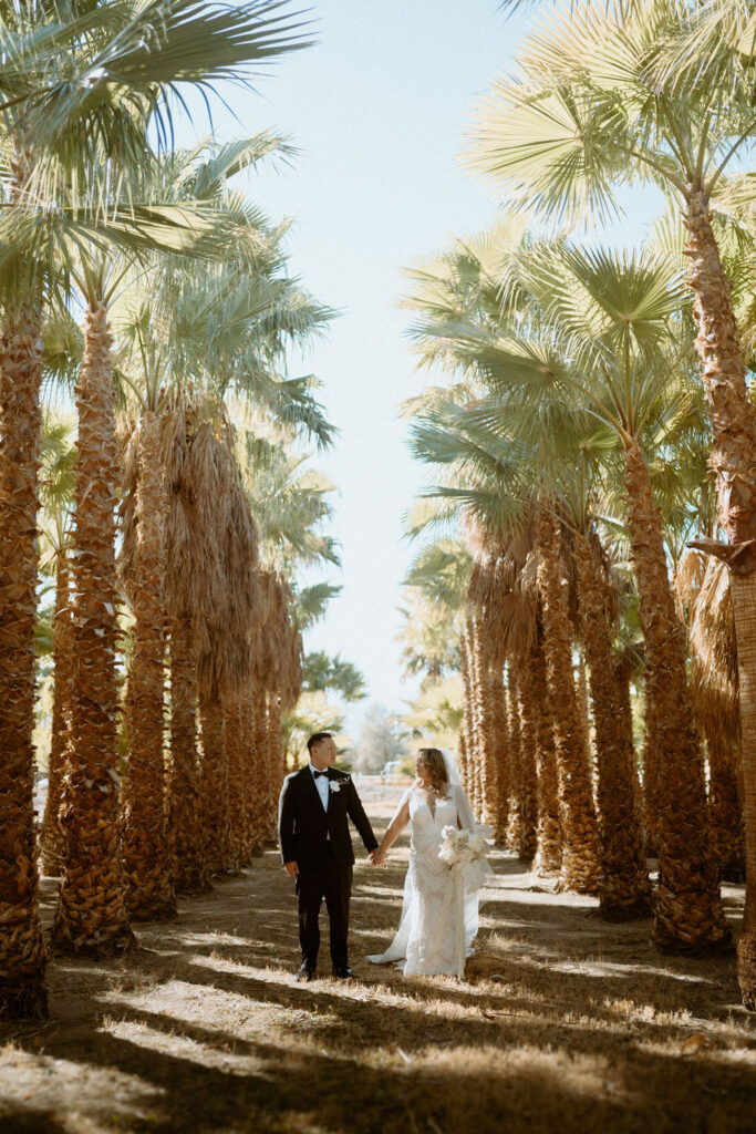 Bride and groom holding hands walking through palm trees 