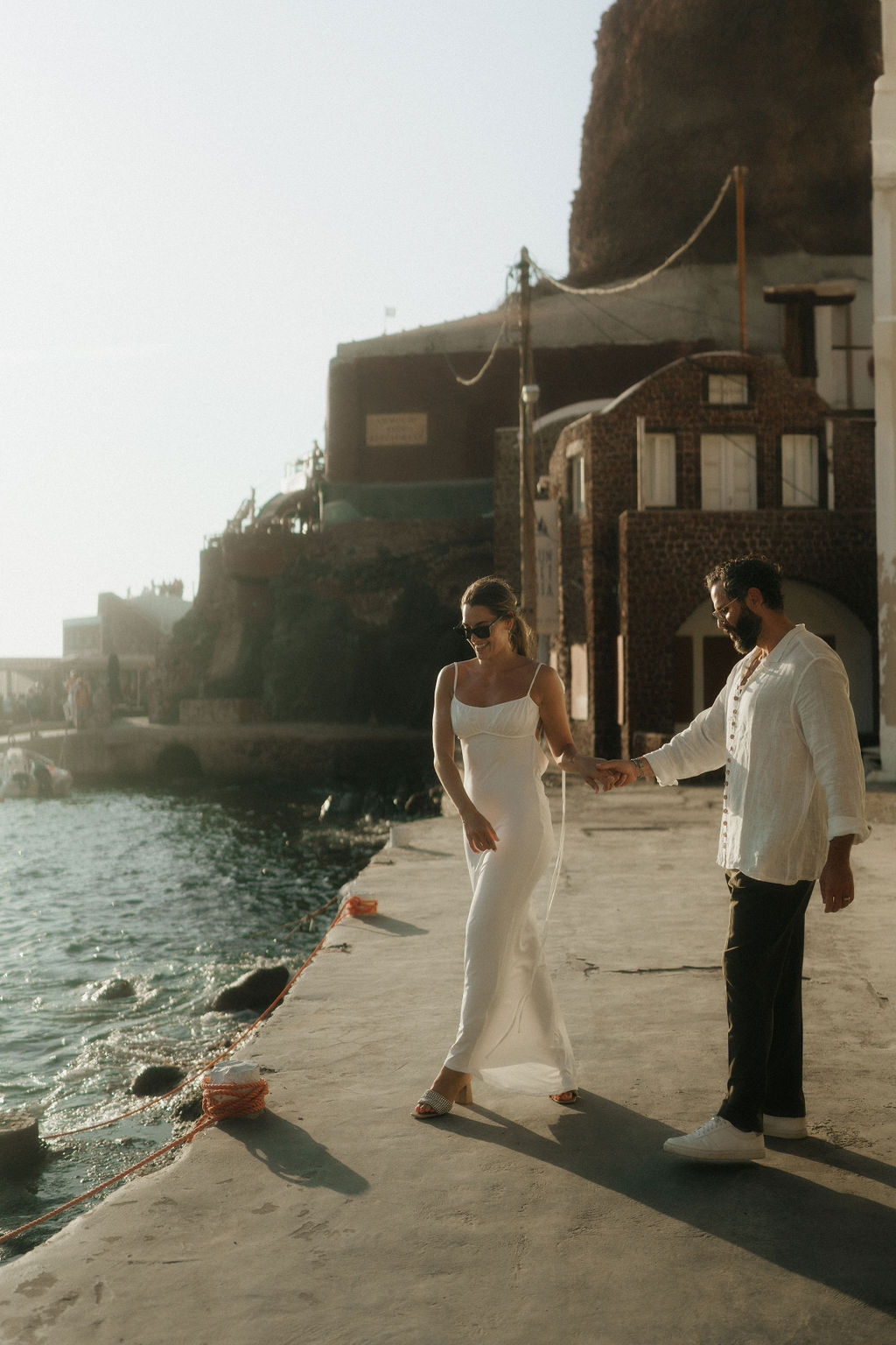 A couple walking hand in hand along a seaside promenade, with the woman wearing a white dress and the man in a white shirt 