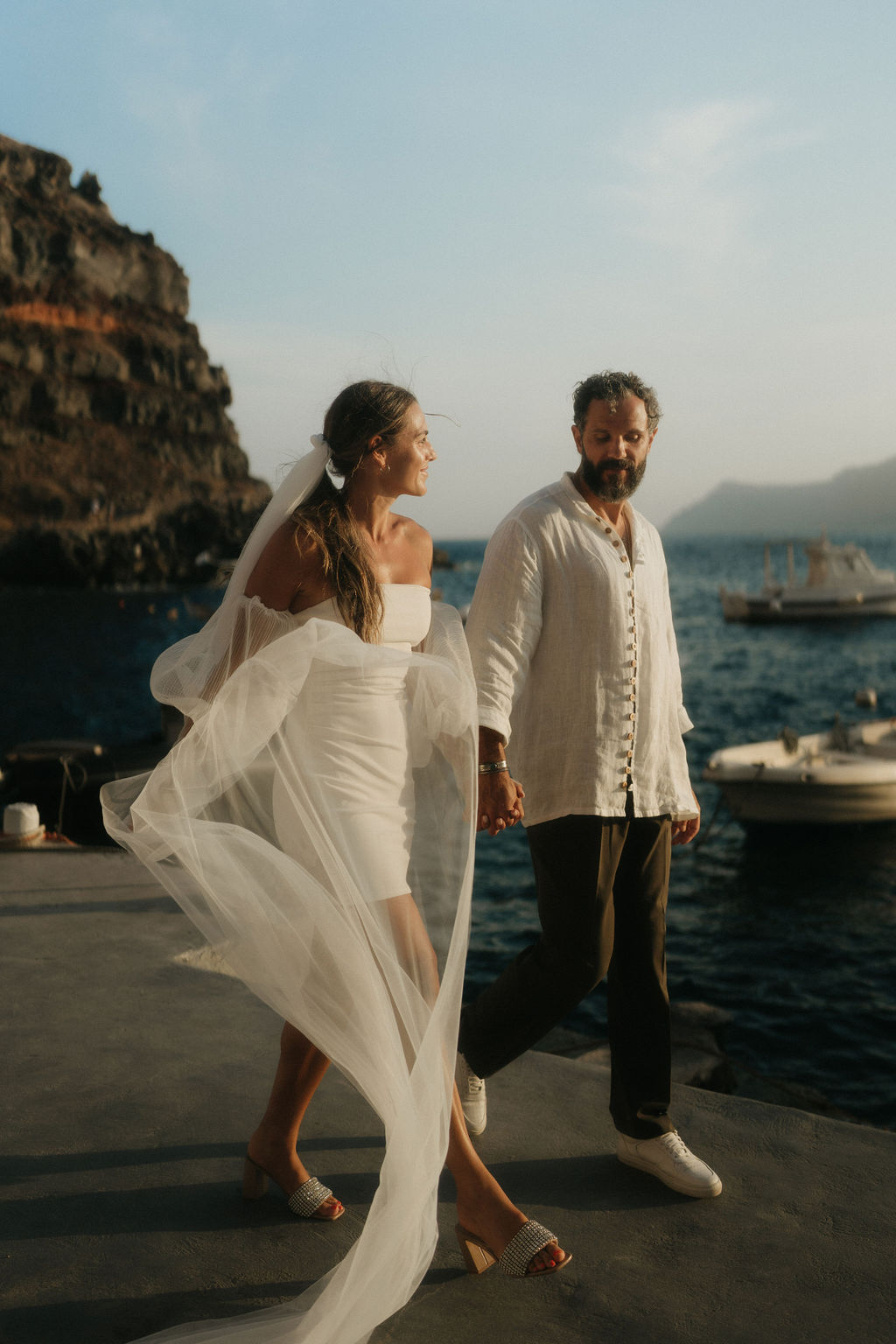 A bride and groom holding hands and walking on a pier, with the sea and hills in the background in santorini greece