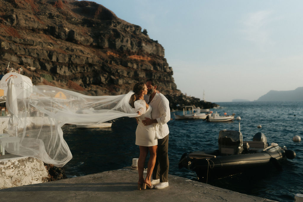 A couple kissing along a seaside, with the woman wearing a white dress and the man in a white shirt 
