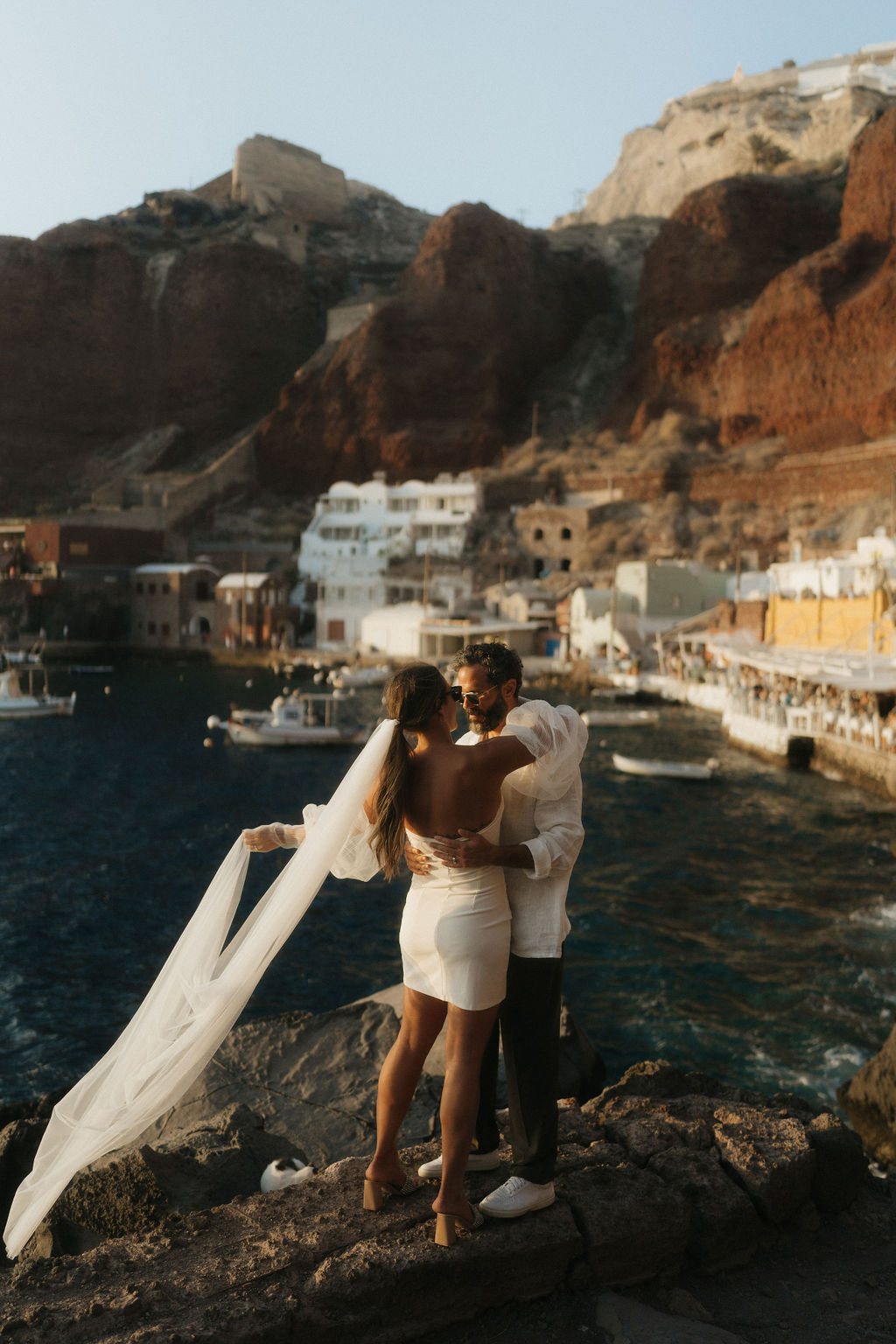 A couple embraces on a rocky shore with a scenic view of the sea and cliffside buildings in the background at their after wedding session in Santorini