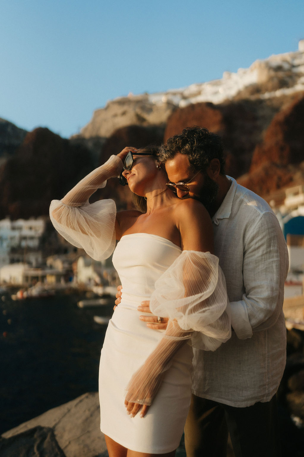 A couple embraces on a rocky shore with a scenic view of the sea and cliffside buildings in the background at their after wedding session in Santorini