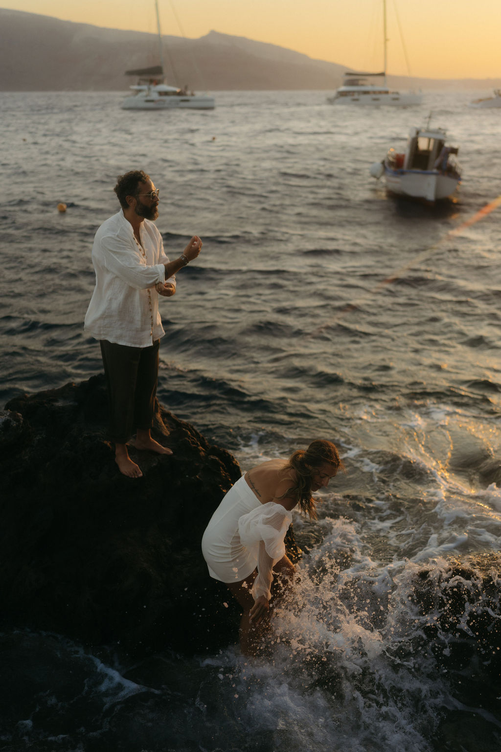 Two people by the sea at dusk, one standing on rocks and another with water splashing around them at their after wedding session 