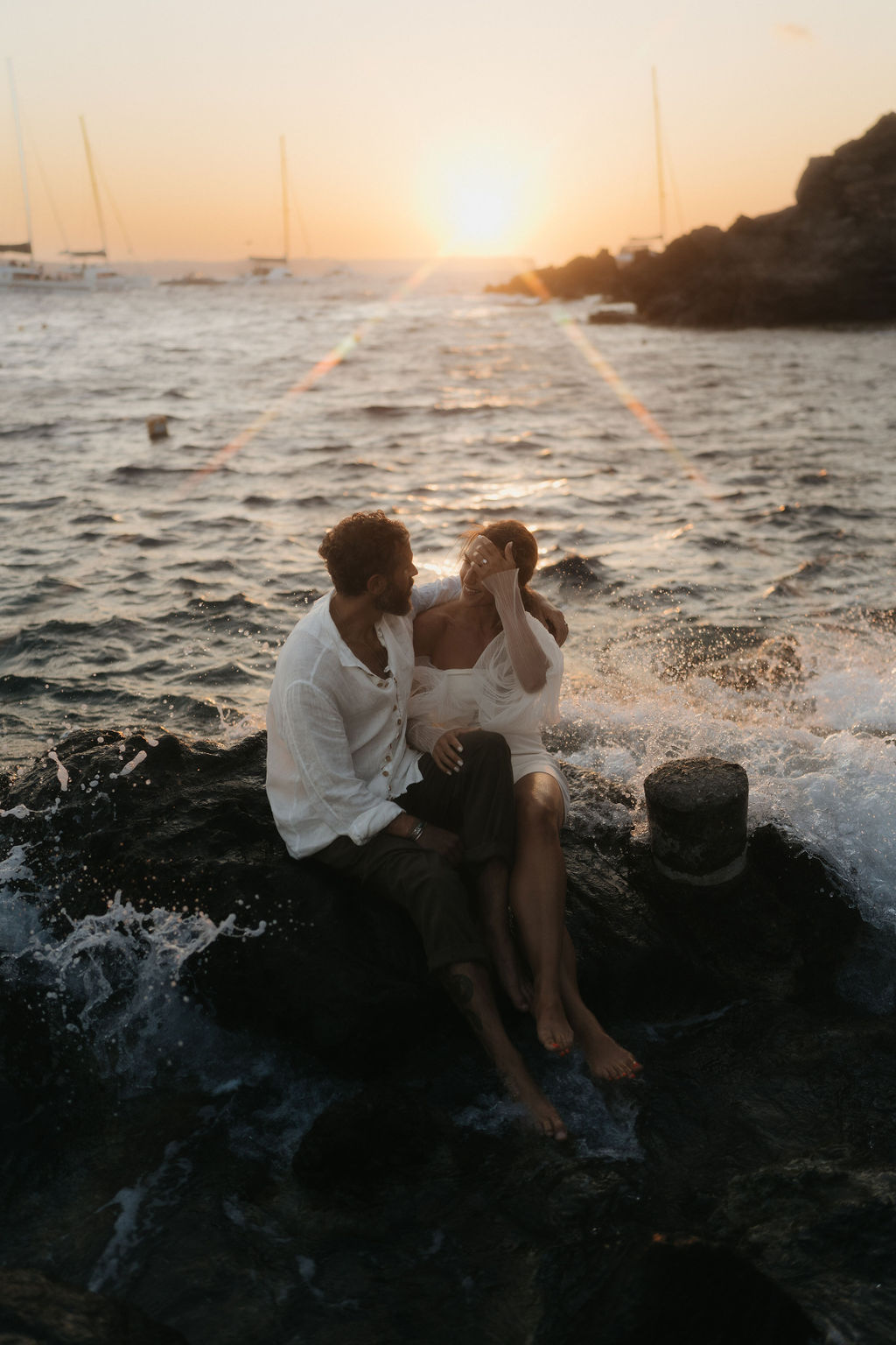 A couple sitting on a rock by the sea, sharing an intimate moment at sunset with waves crashing nearby at their after wedding session 