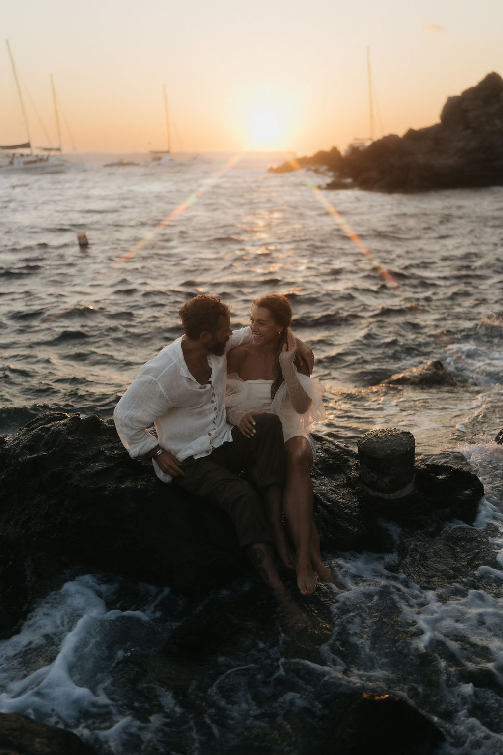 A couple sitting on a rock by the sea, sharing an intimate moment at sunset with waves crashing nearby at their after wedding session