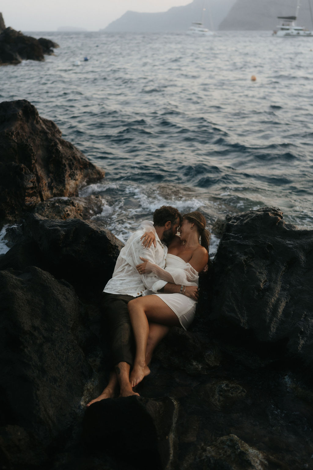 A couple sitting on a rock by the sea, sharing an intimate moment at sunset with waves crashing nearby at their after wedding session 