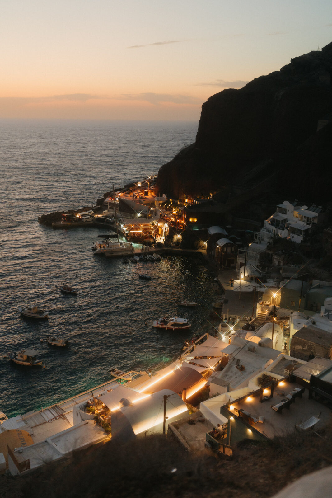 Santorini Greece at dusk, with boats floating near the shore and a sunset over the horizon.