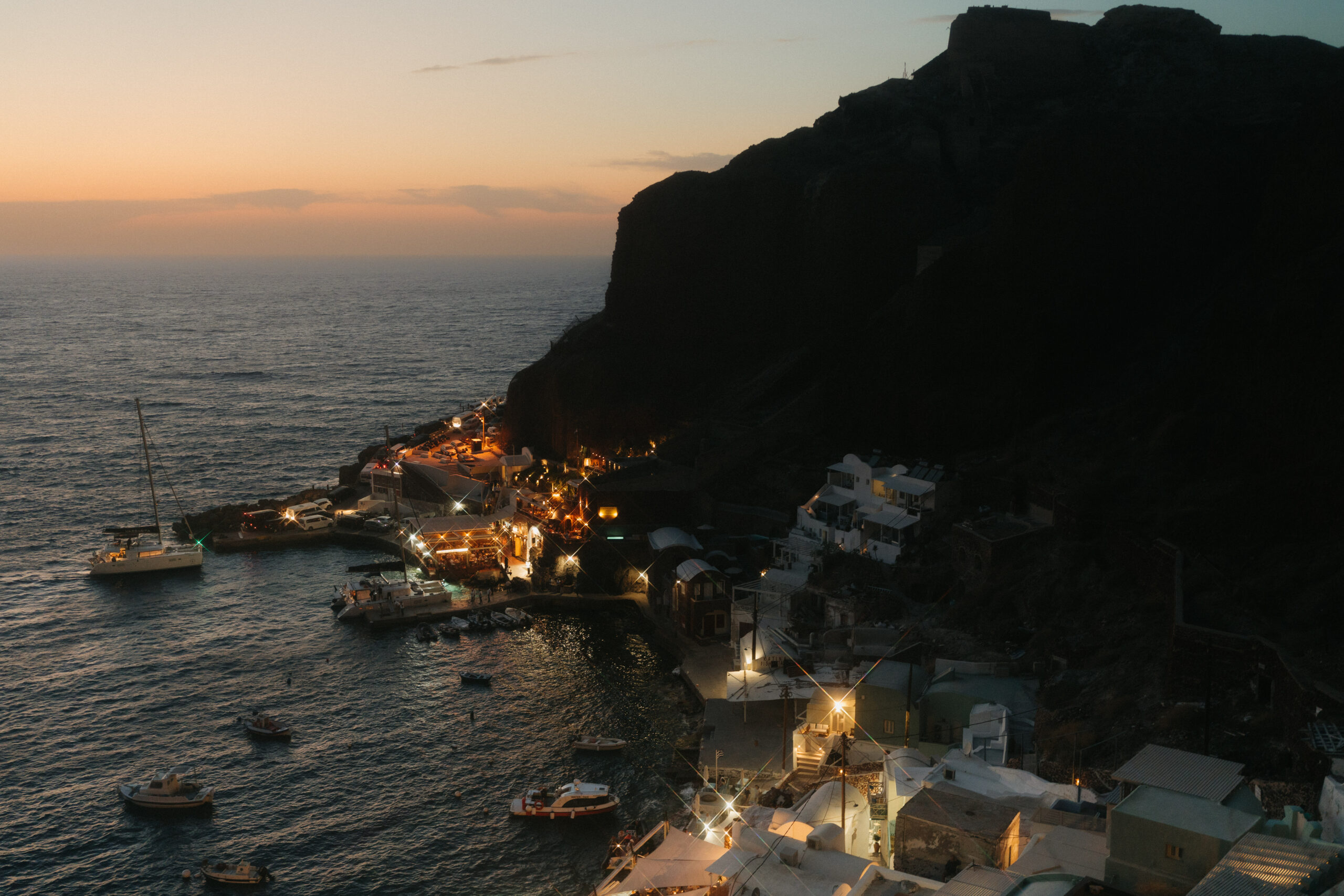 Santorini Greece at dusk, with boats floating near the shore and a sunset over the horizon.