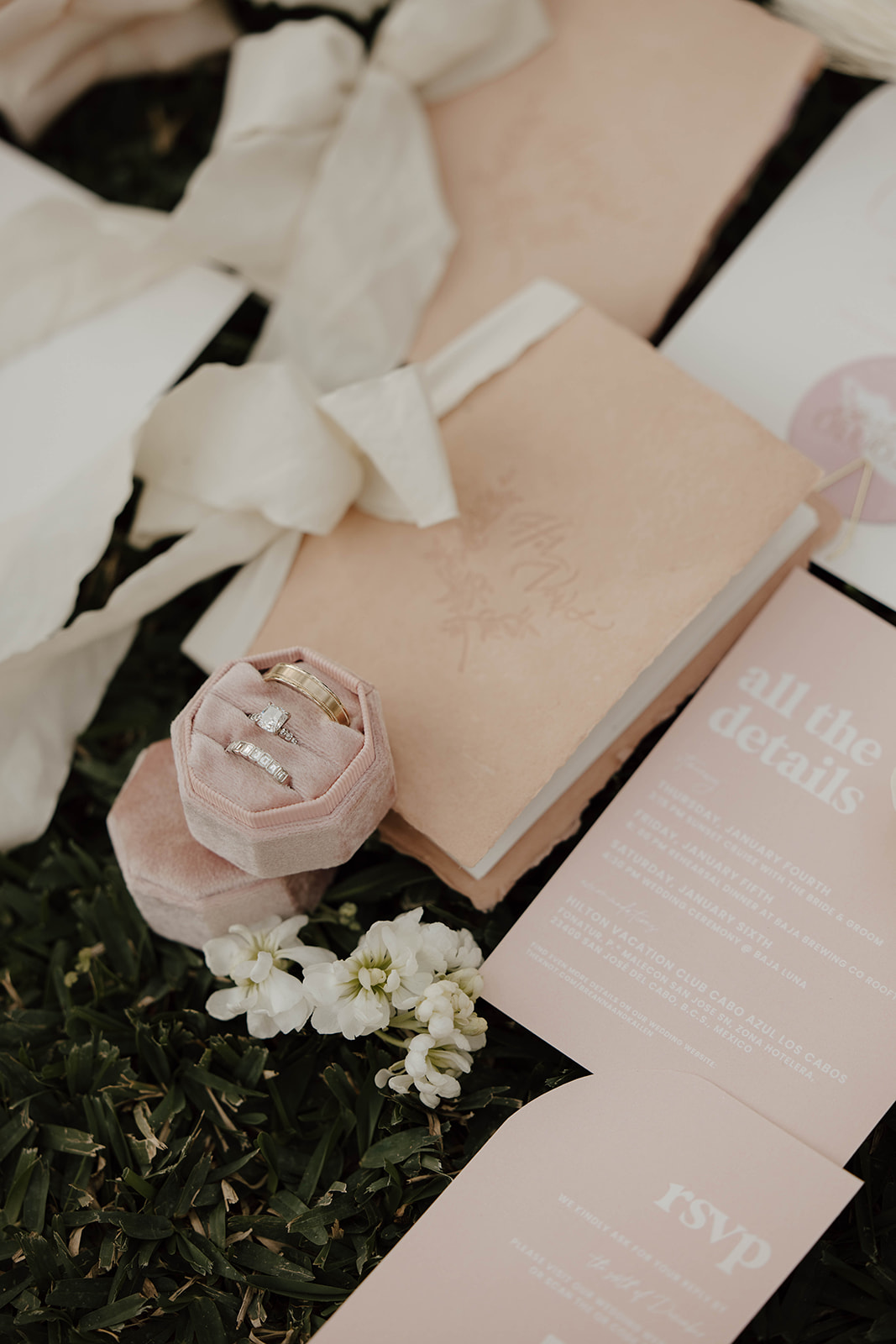 An elegant wedding invitation suite with a ring nestled in a soft pink fabric, accompanied by white florals on a grassy background.