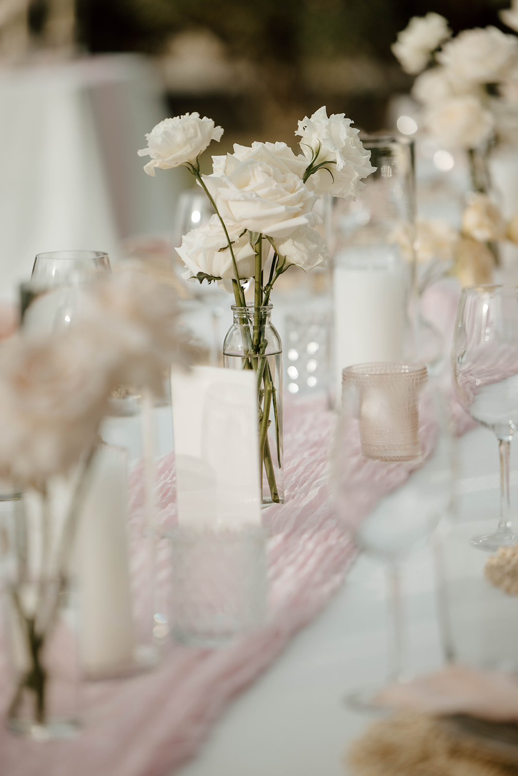 Elegant table setting with white flowers in a glass vase, soft pink textiles, and delicate glassware.