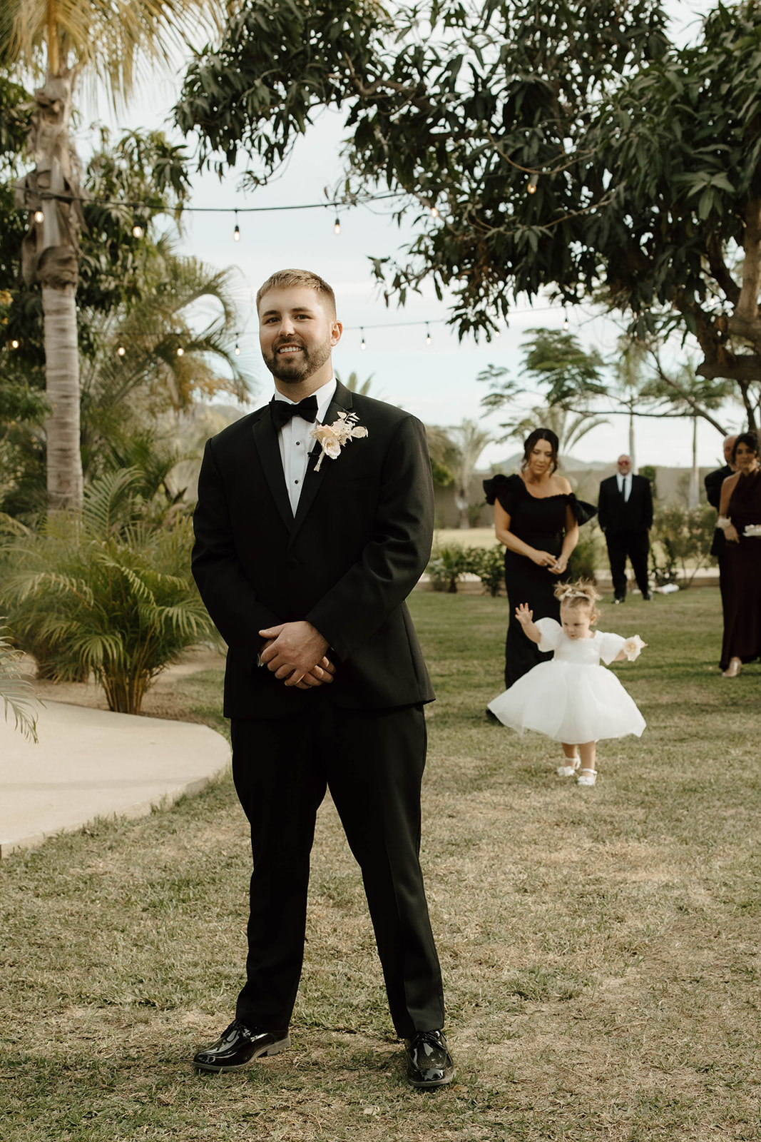 A man in a black tuxedo stands smiling at an outdoor wedding, while a child in a white dress walks behind and guests look on to do a father- daughter first look
