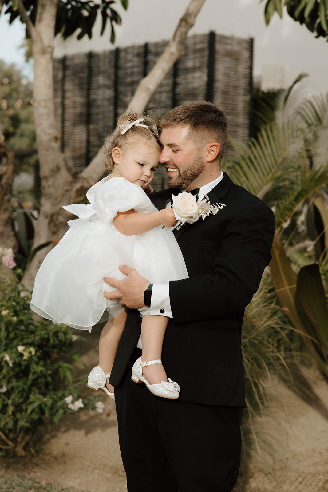 Groom in a black suit holding a smiling young girl in a white dress at a Cabo San Lucas wedding