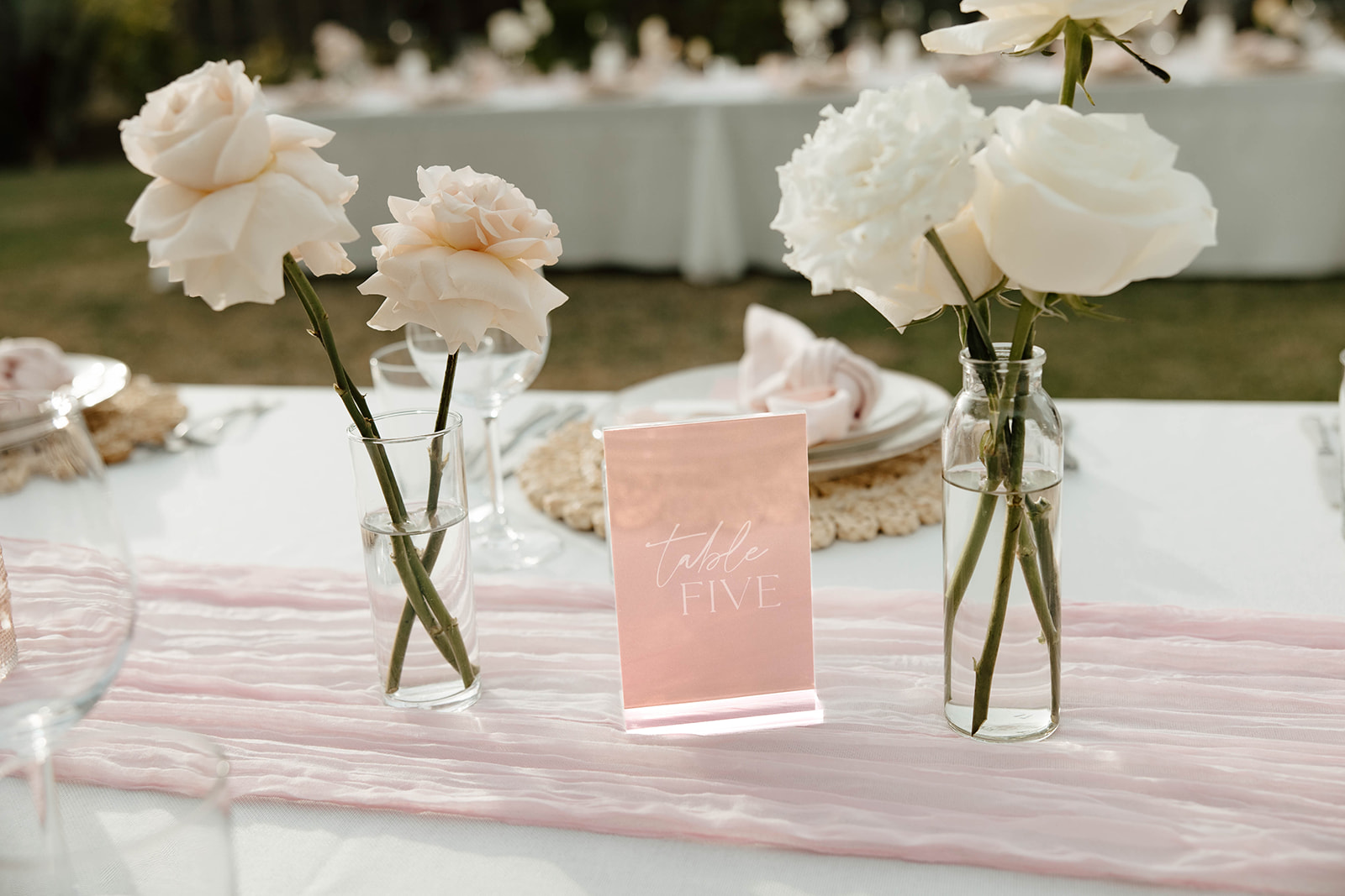 Elegant outdoor table setting with a "table five" sign, pale pink table runner, and white roses in glass vases for a cabo san lucas wedding