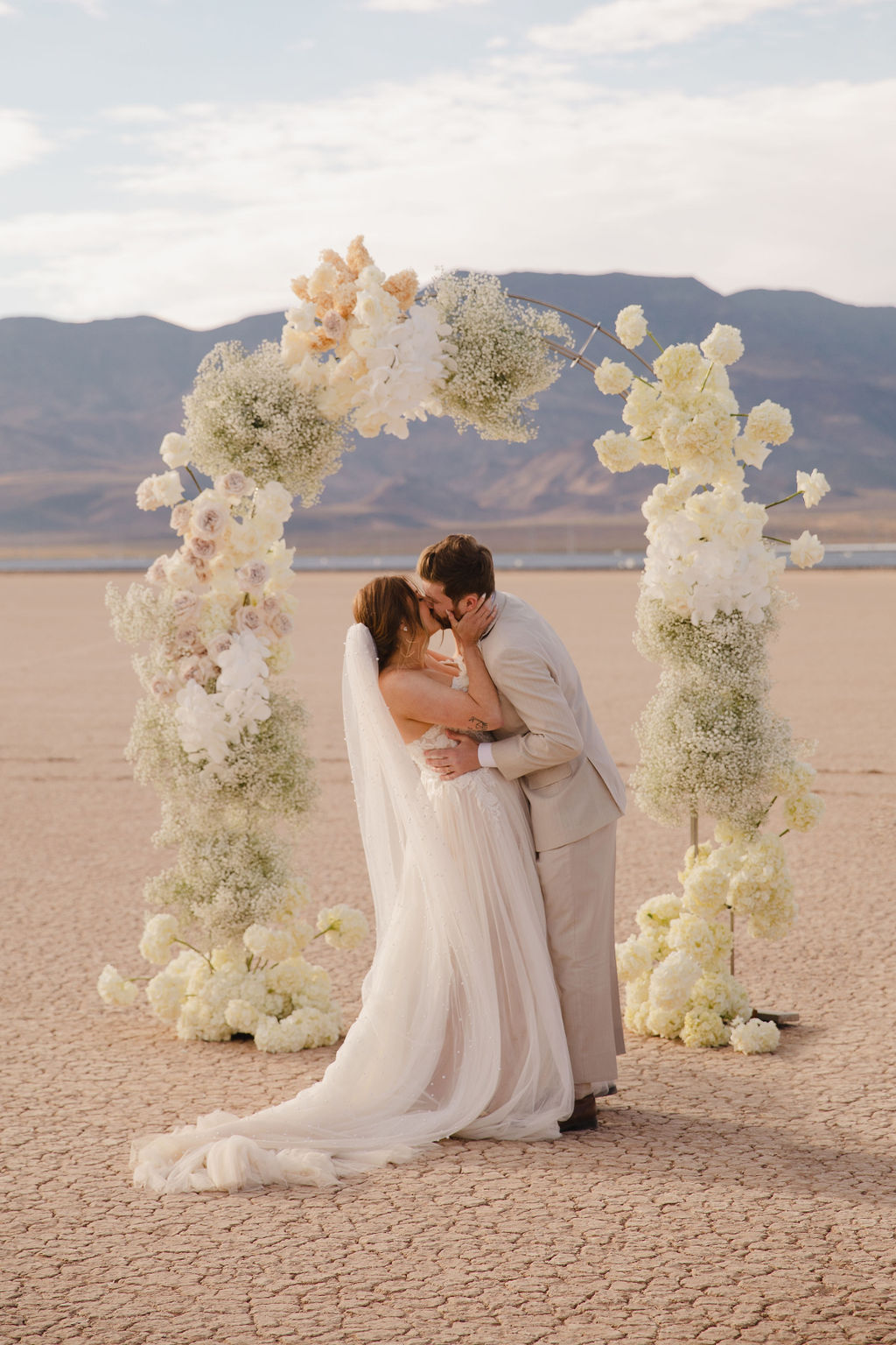 A couple holds hands during a wedding ceremony in a desert setting, officiated by a woman, under a floral arch at their Dry Lake Bed wedding