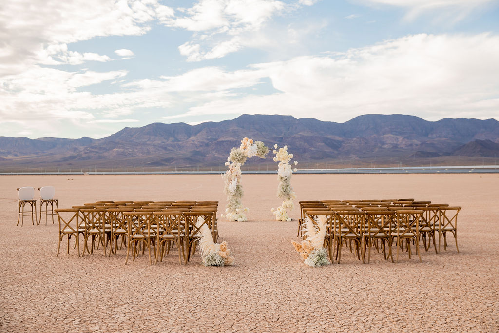 Outdoor wedding setup in a desert with rows of wooden chairs and a floral arch, mountains in the background under a cloudy sky.