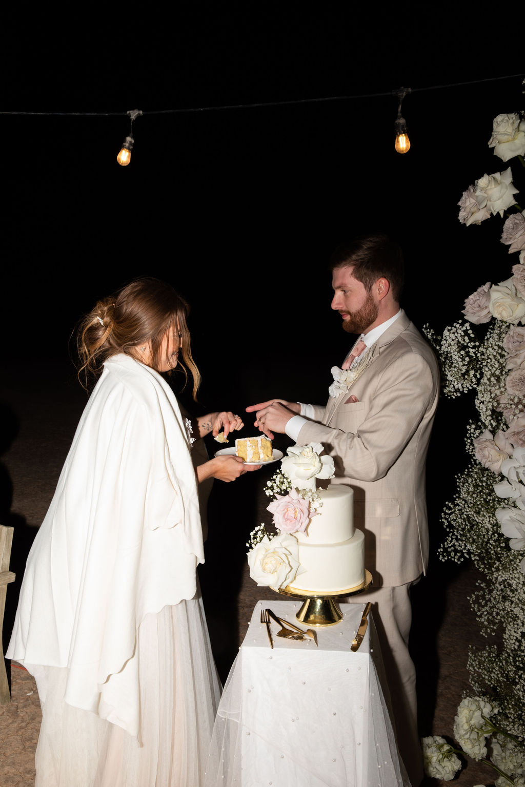 A bride and groom cut their wedding cake at night under string lights, with a guest sitting nearby.