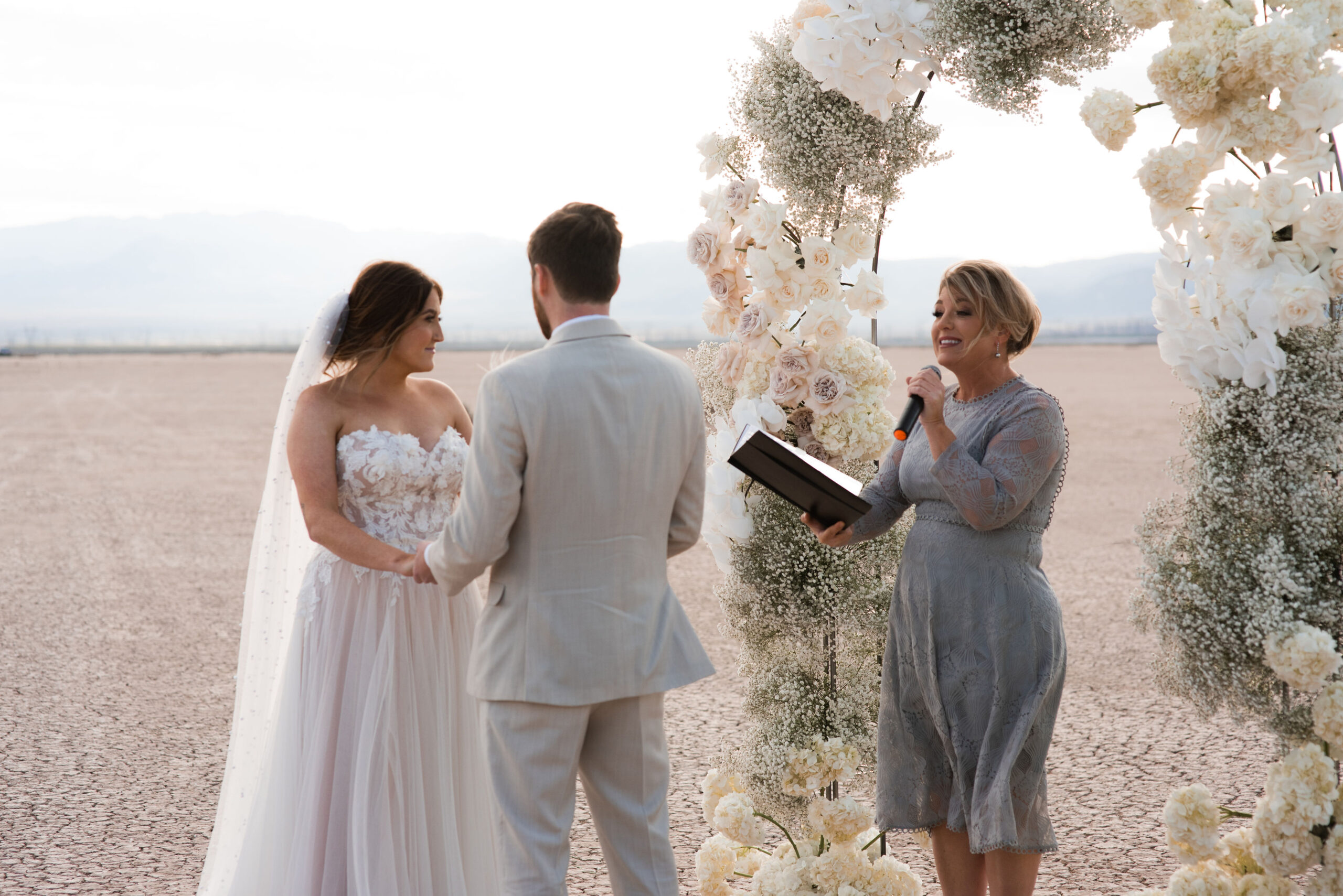A couple holds hands during a wedding ceremony in a desert setting, officiated by a woman, under a floral arch at their Dry Lake Bed wedding