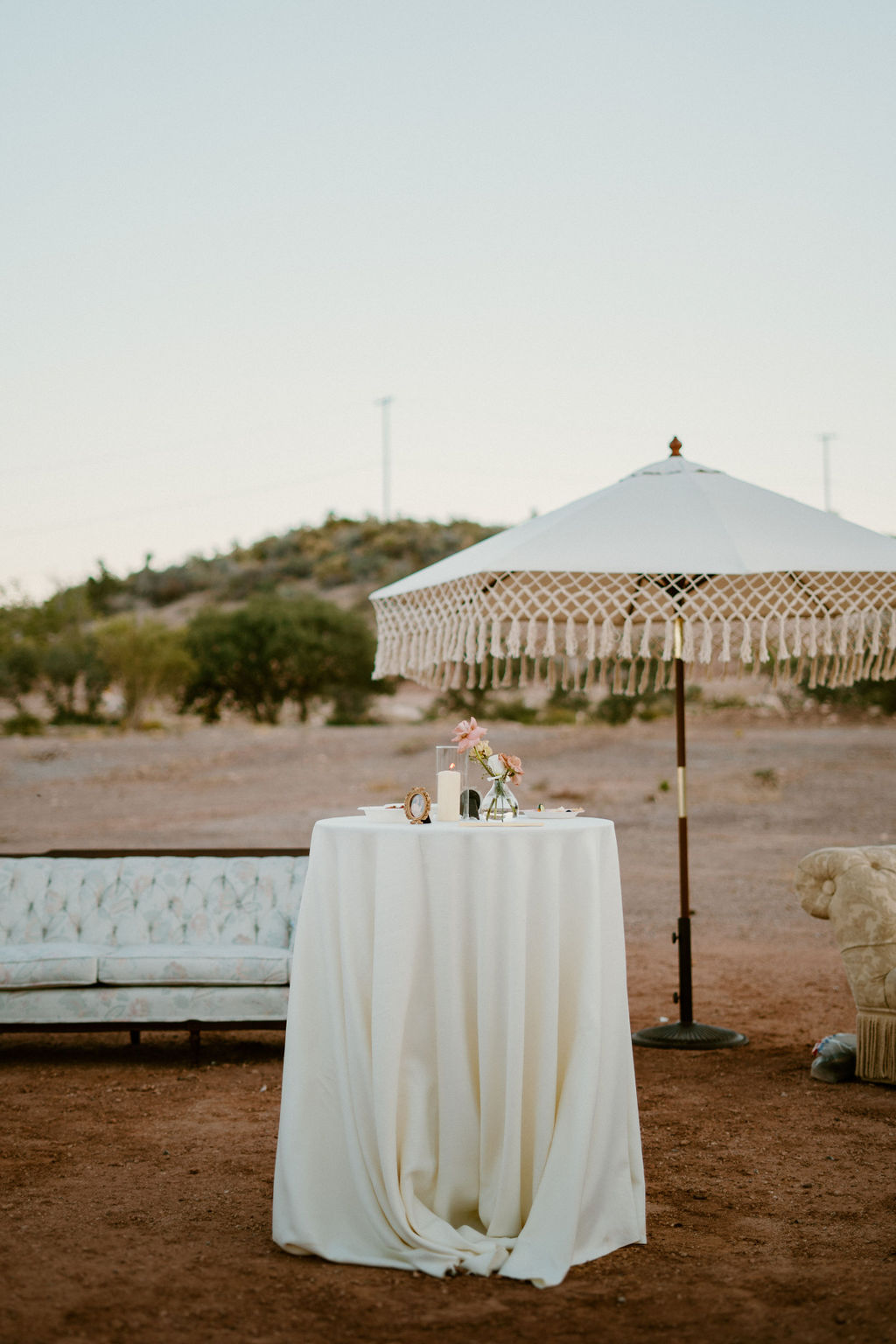 An outdoor event setup featuring a small round table with a white cloth, decorative items on top, and a large umbrella, surrounded by a natural landscape and a vintage sofa.