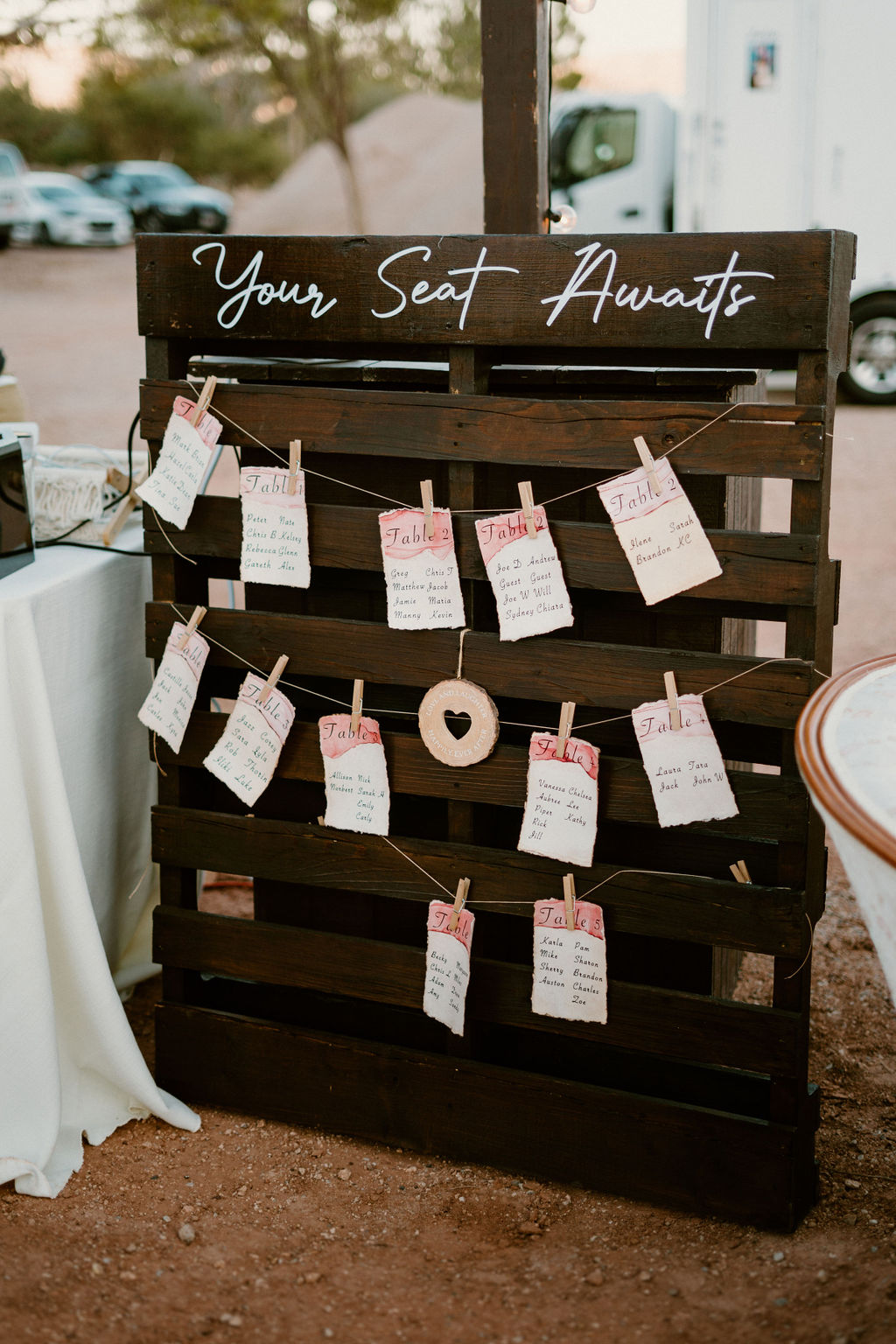 Rustic wooden seating chart with cards hanging, labeled "your seat awaits," at an outdoor event.