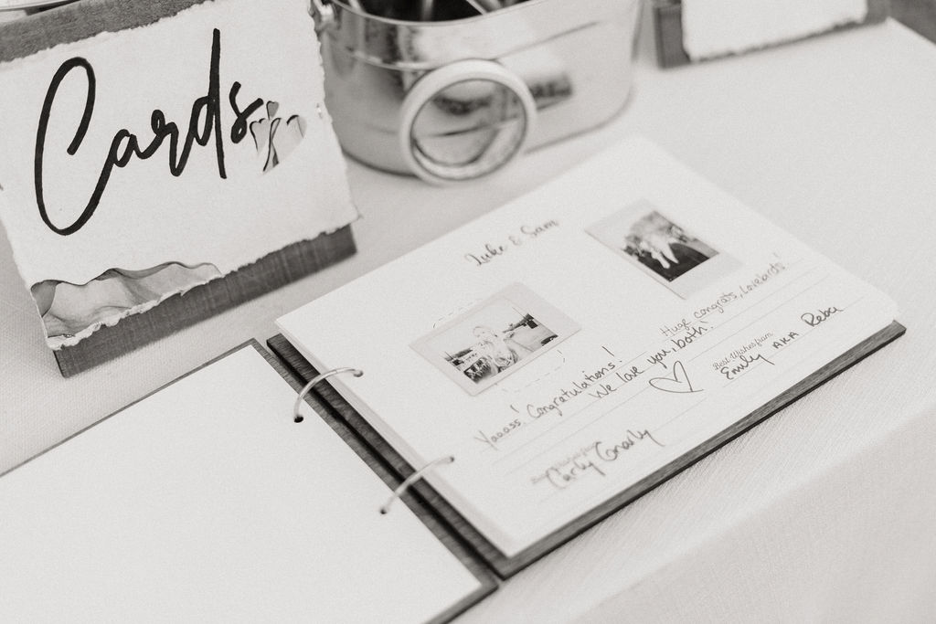 Black and white photo of a guest book open to a page with handwritten messages and photos, next to a sign reading "cards" and a metal bucket.