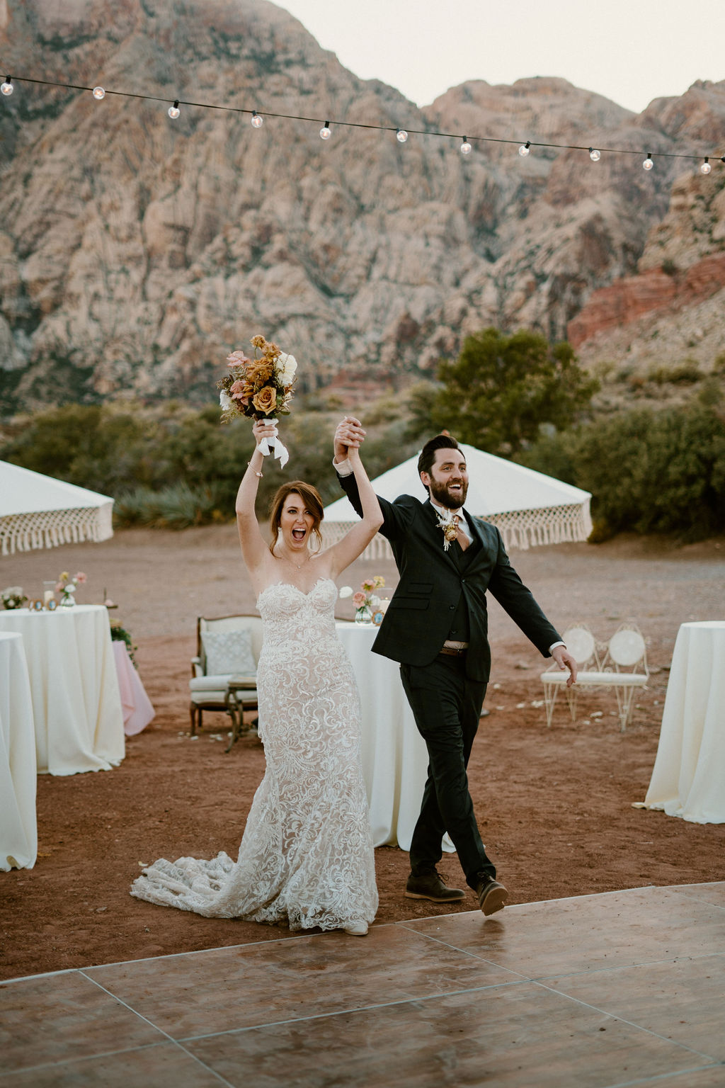 A bride and groom walk hand in hand at an outdoor wedding reception with tables, decorative umbrellas, and string lights, with a dramatic mountain backdrop at Red Rock Canyon