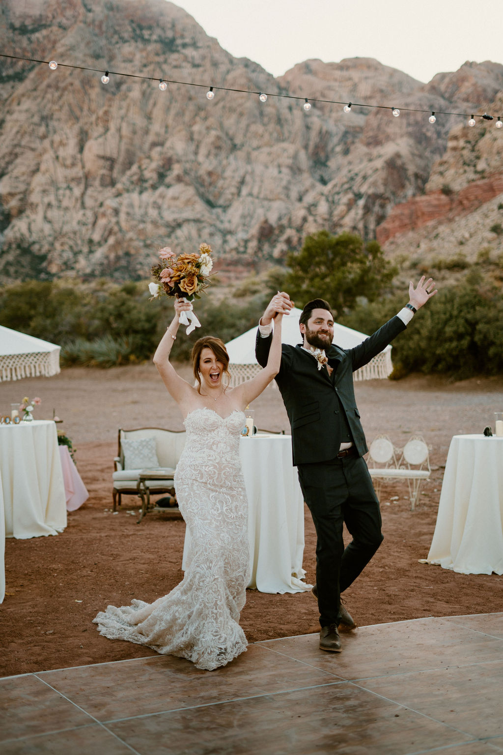 A bride and groom walk hand in hand at an outdoor wedding reception with tables, decorative umbrellas, and string lights, with a dramatic mountain backdrop at Red Rock Canyon