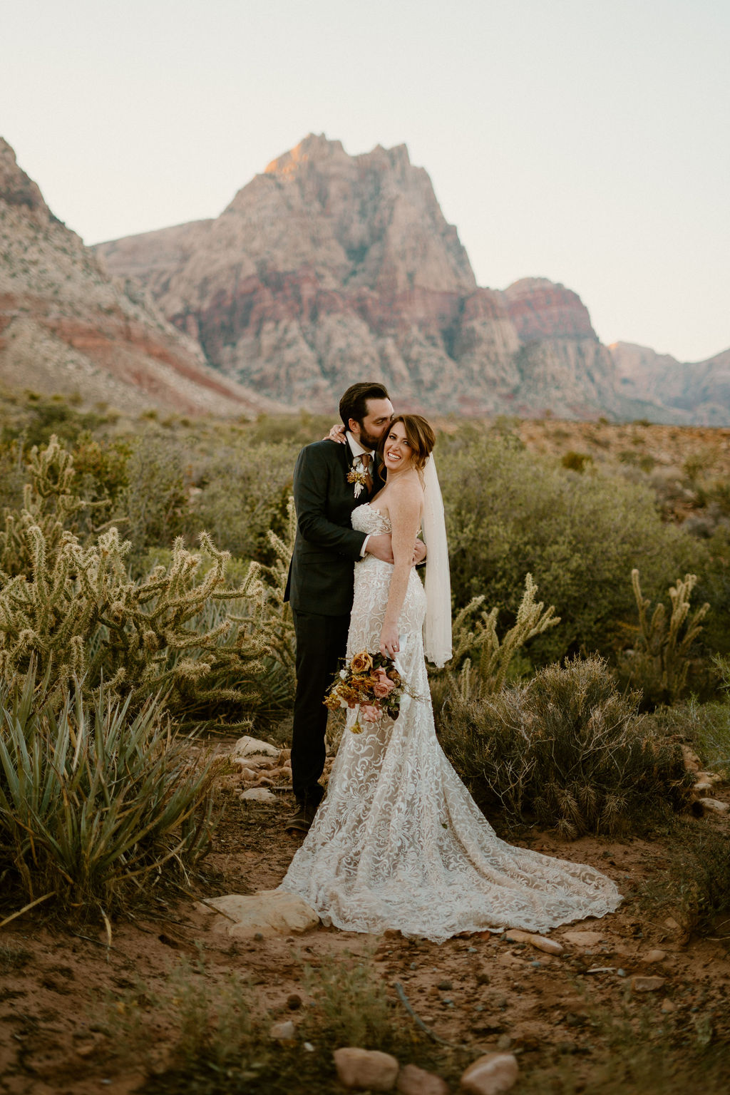 A bride and groom smiling and embracing in a desert landscape with rocky mountains in the background.