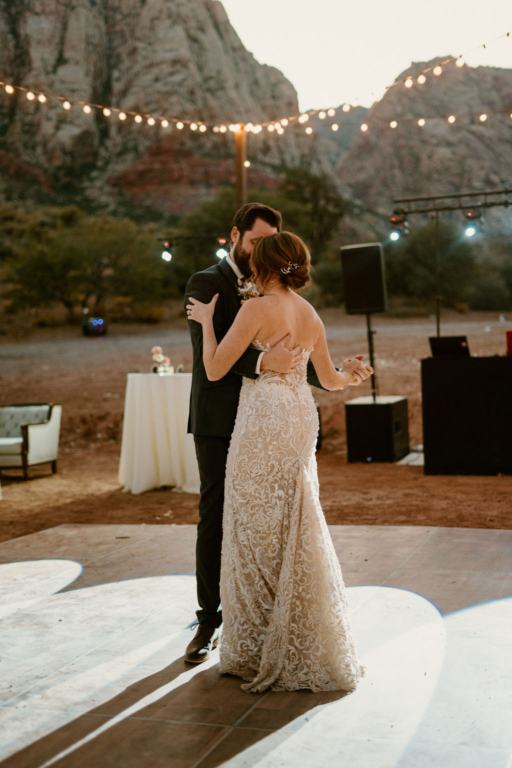 A couple in formal attire sharing a dance outdoors at sunset, with string lights and mountains in the background.