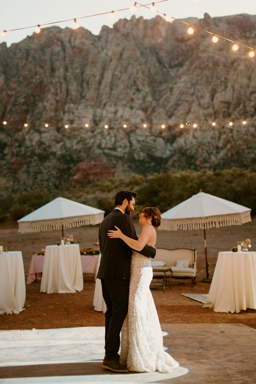 A couple in formal attire sharing a dance outdoors at sunset, with string lights and mountains in the background.