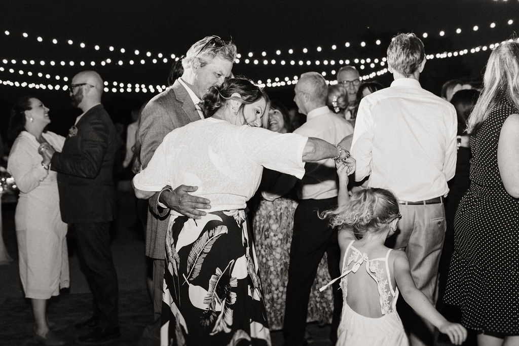 A couple dances intimately under string lights at a nighttime outdoor event, surrounded by other dancing couples.