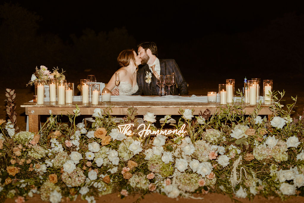 A couple kisses at a candlelit table adorned with flowers in a nighttime setting, a sign reading "the johnsons" visible in front.