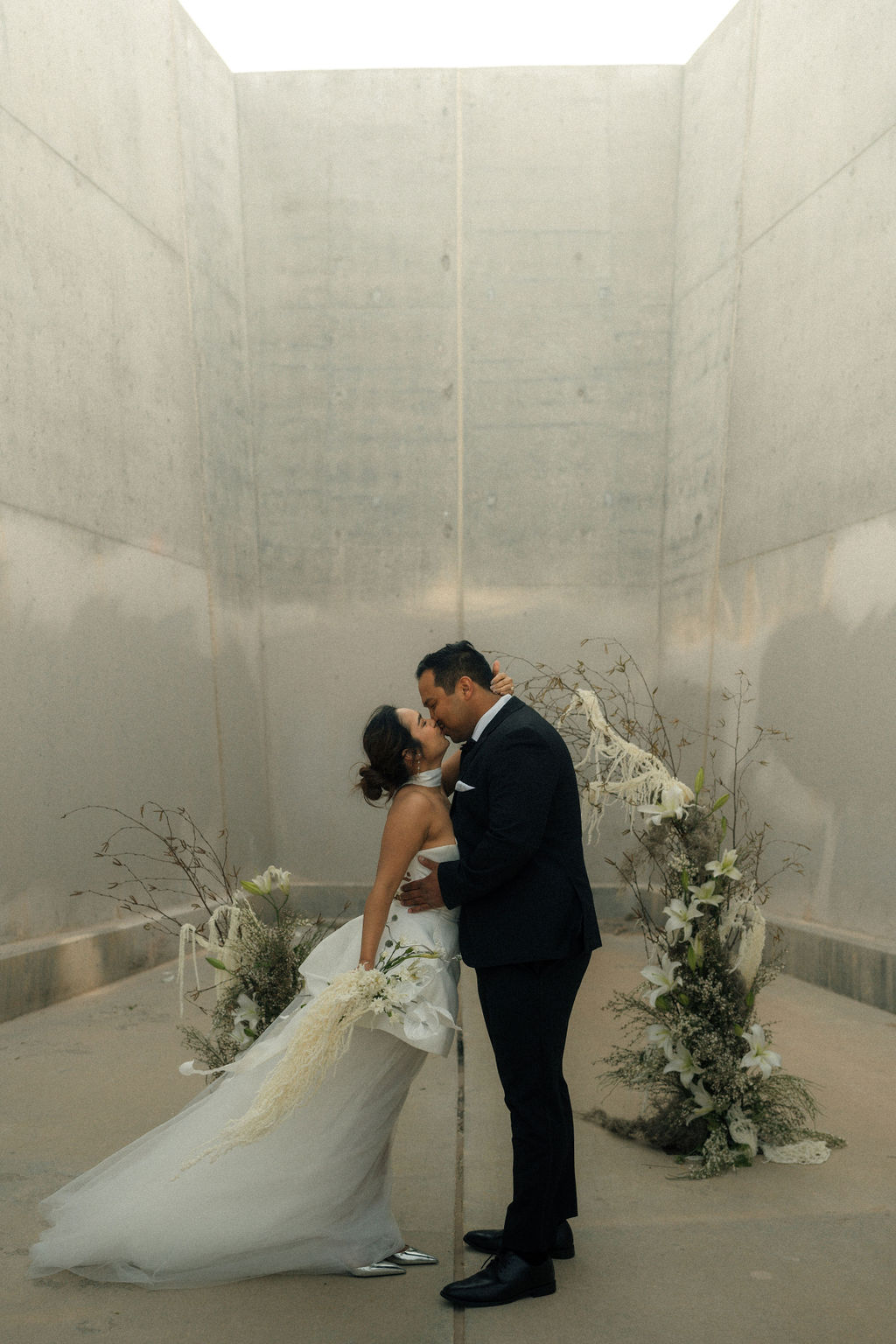 A bride and groom share a kiss in a minimalist concrete corridor, surrounded by large floral arrangements at their intimate wedding