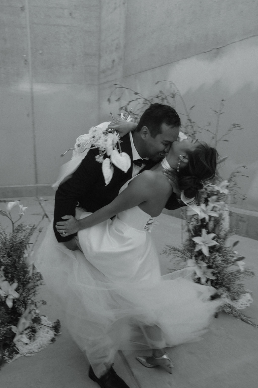 A bride and groom share a kiss in a minimalist concrete corridor, surrounded by large floral arrangements at their intimate wedding